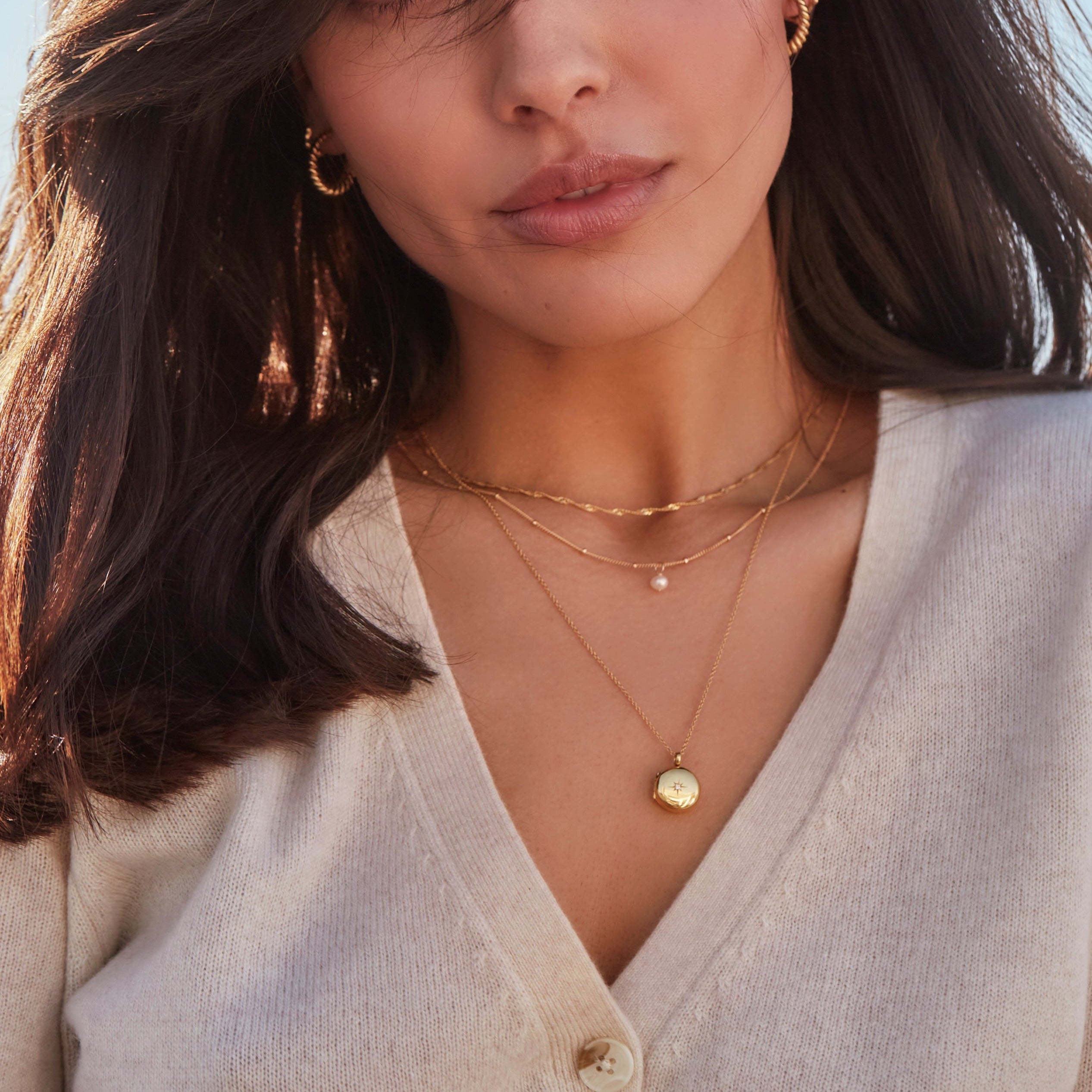 Woman wearing a gold small round diamond locket necklace, a gold twisted rope chain necklace and a gold single pearl satellite necklace around her neck with a beige jumper