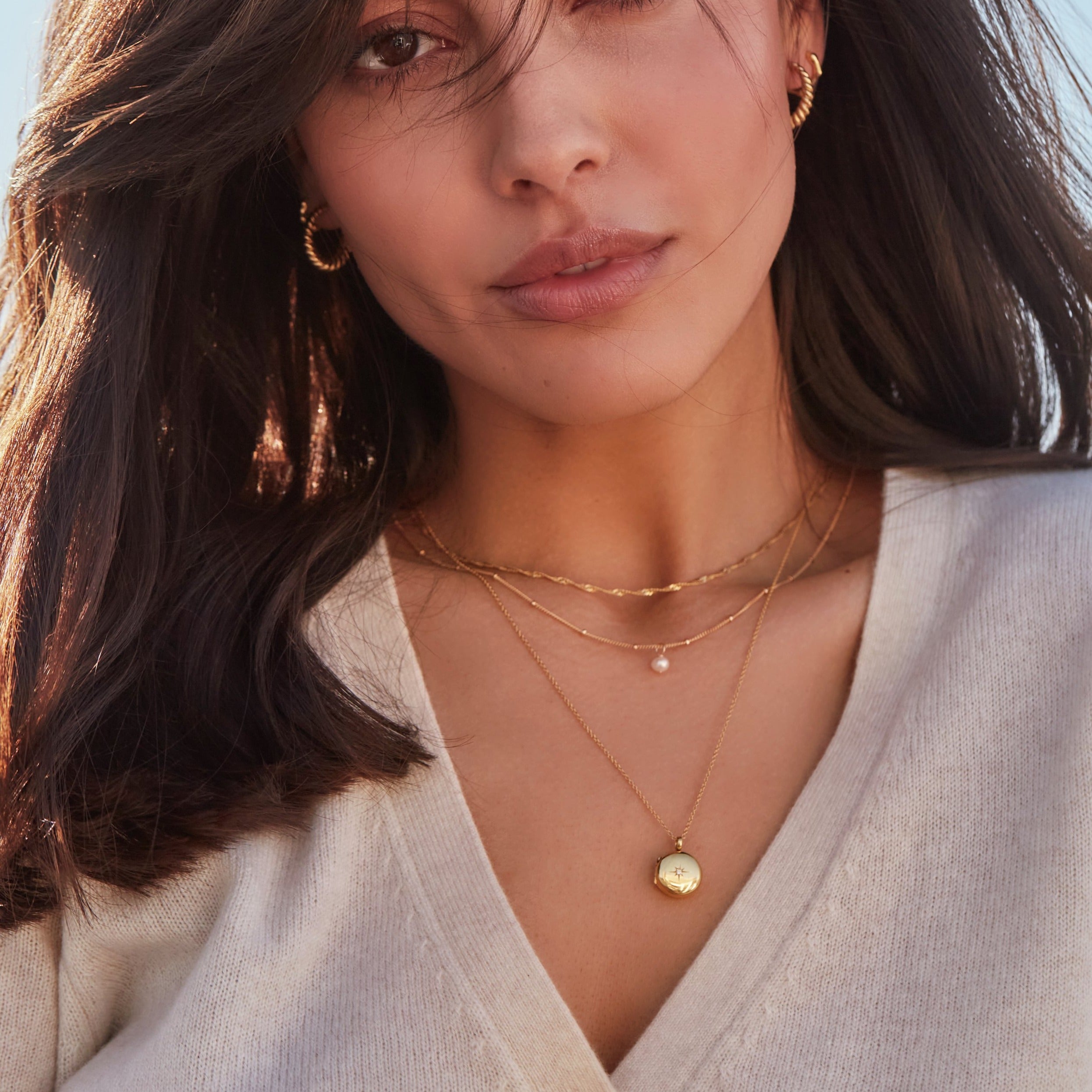 Brunette woman wearing a gold single pearl satellite necklace layered with a gold twisted rope necklace and gold small round diamond locket necklace with gold large twisted rope hoop earrings in her ears 