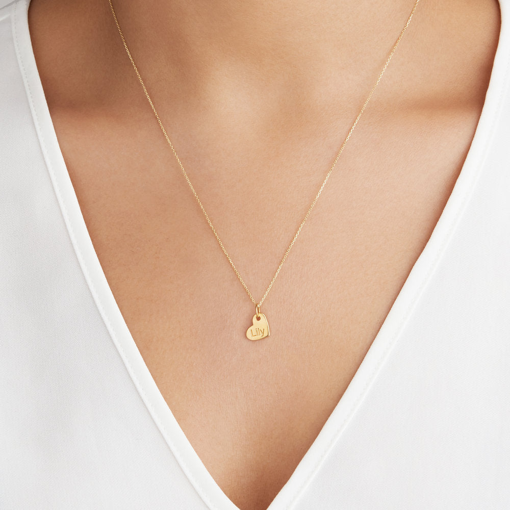 Gold small personalised heart necklace with the name 'Lily' engraved on a chest of a woman wearing a white V-neck top