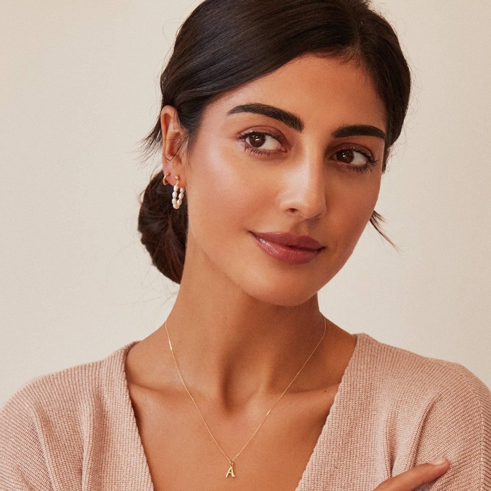 Gold seed pearl hoop earring in ear lobe of a dark haired woman looking to the side with a silver individual curve initial charm 'A' around her neck