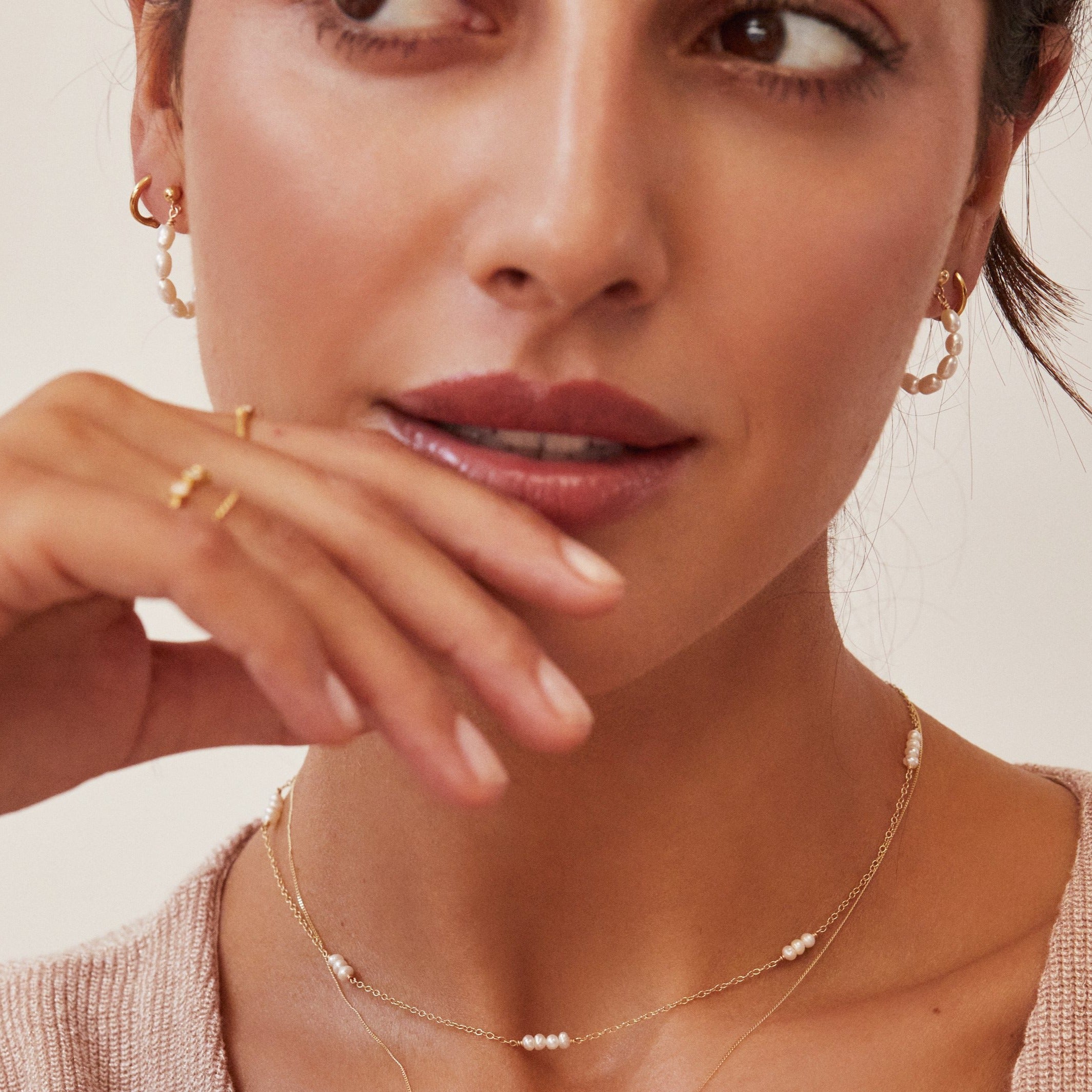 A woman wearing gold seed pearl hoop earrings and a pearl necklace with her eyes looking to the side