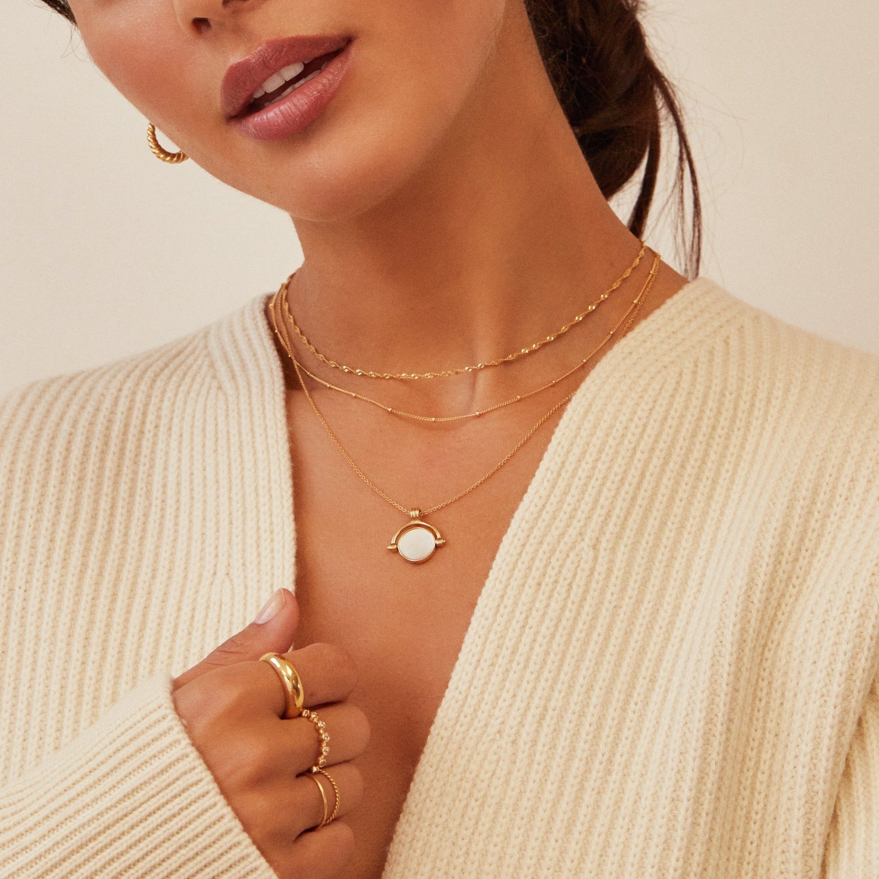 Close up of brunette woman wearing a gold mother of pearl spinning disc necklace layered with a gold satellite chain necklace and a gold twisted rope necklace, gold plaine dome ring and other gold rings on her fingers and a cream knitted jumper