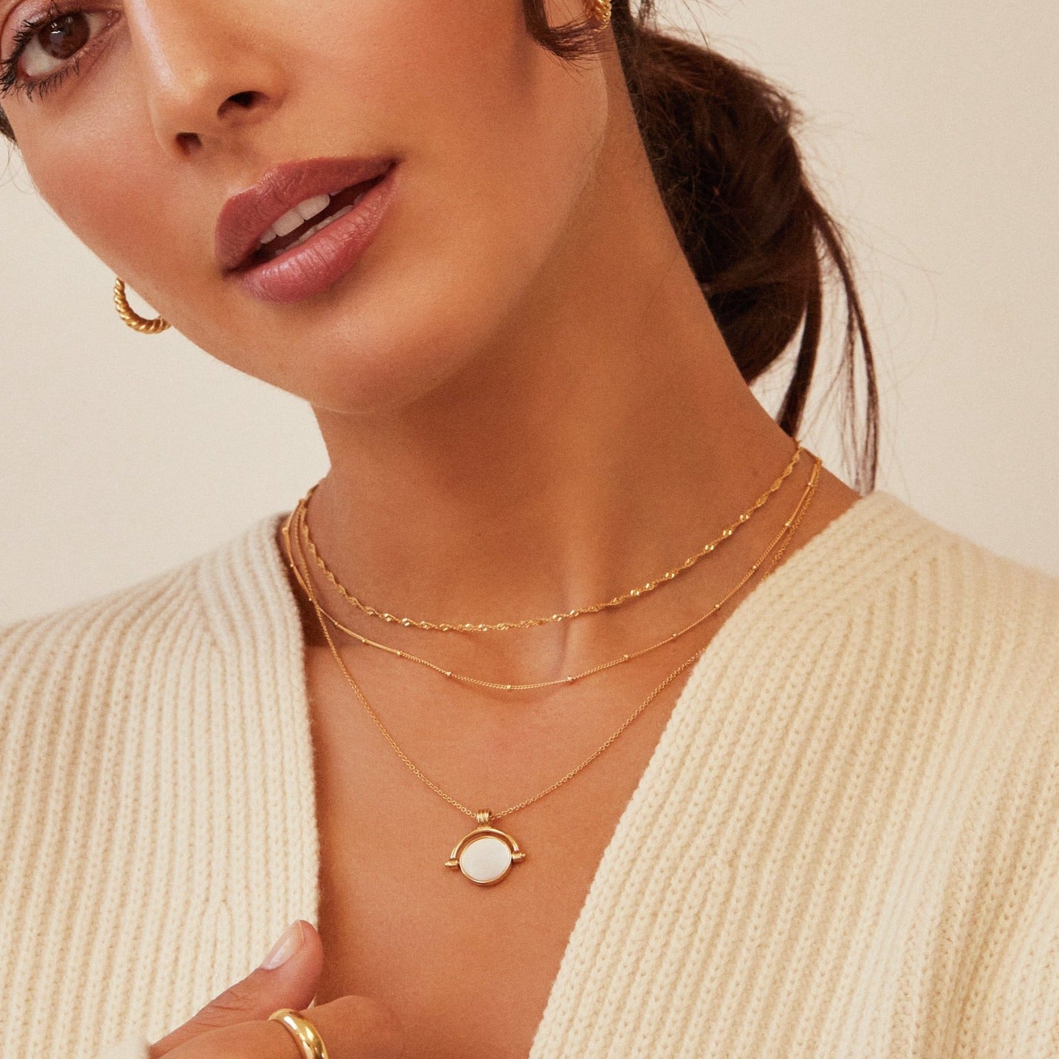 Brunette woman wearing a gold mother of pearl spinning disc necklace layered with a gold satellite chain necklace and a gold twisted rope chain necklace, gold plaine dome ring and other gold rings on her fingers and a cream knitted jumper