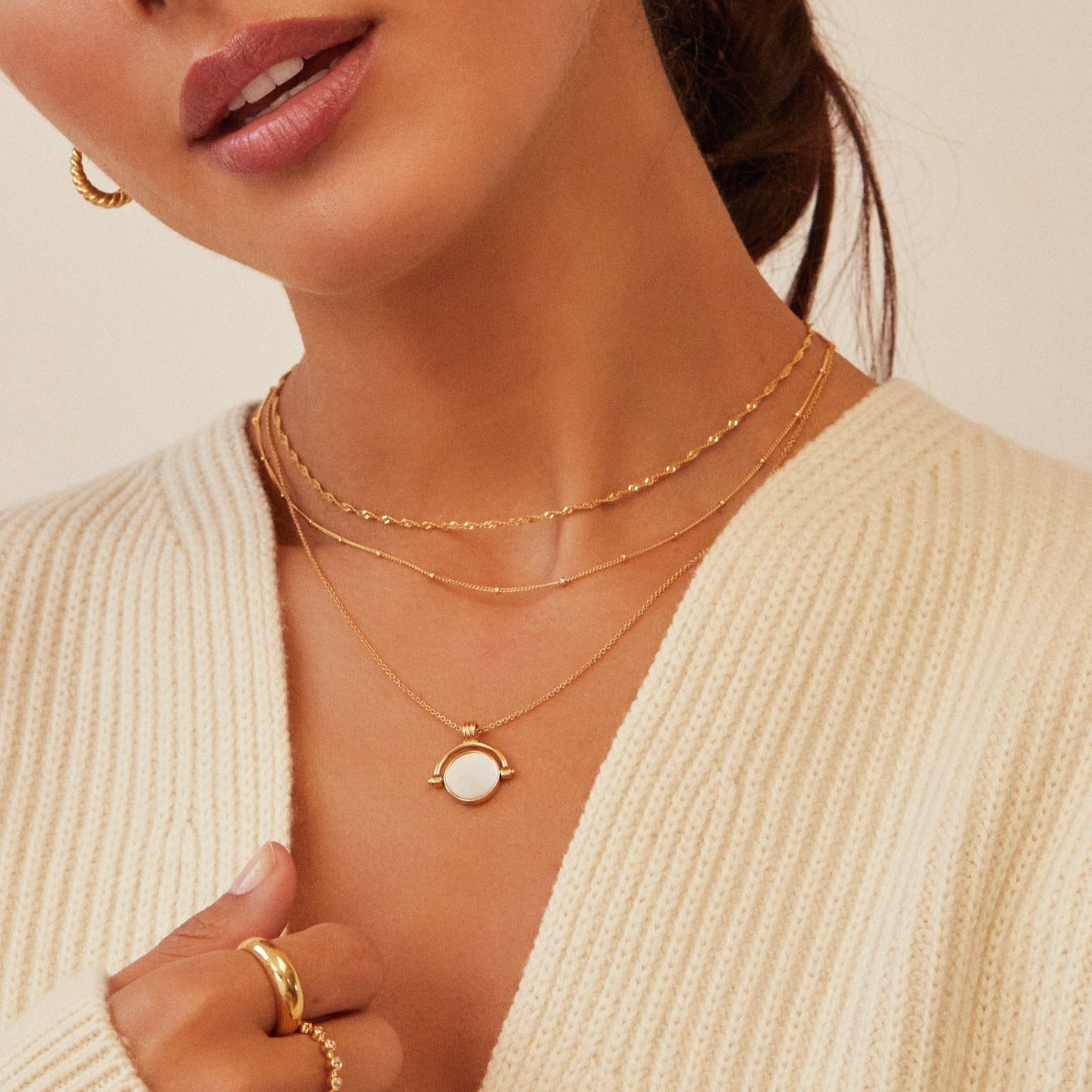 Medium close up of a brunette woman wearing a gold mother of pearl spinning disc necklace layered with a gold satellite chain necklace and a gold twisted rope chain necklace, gold plaine dome ring and other gold rings on her fingers and a cream knitted jumper