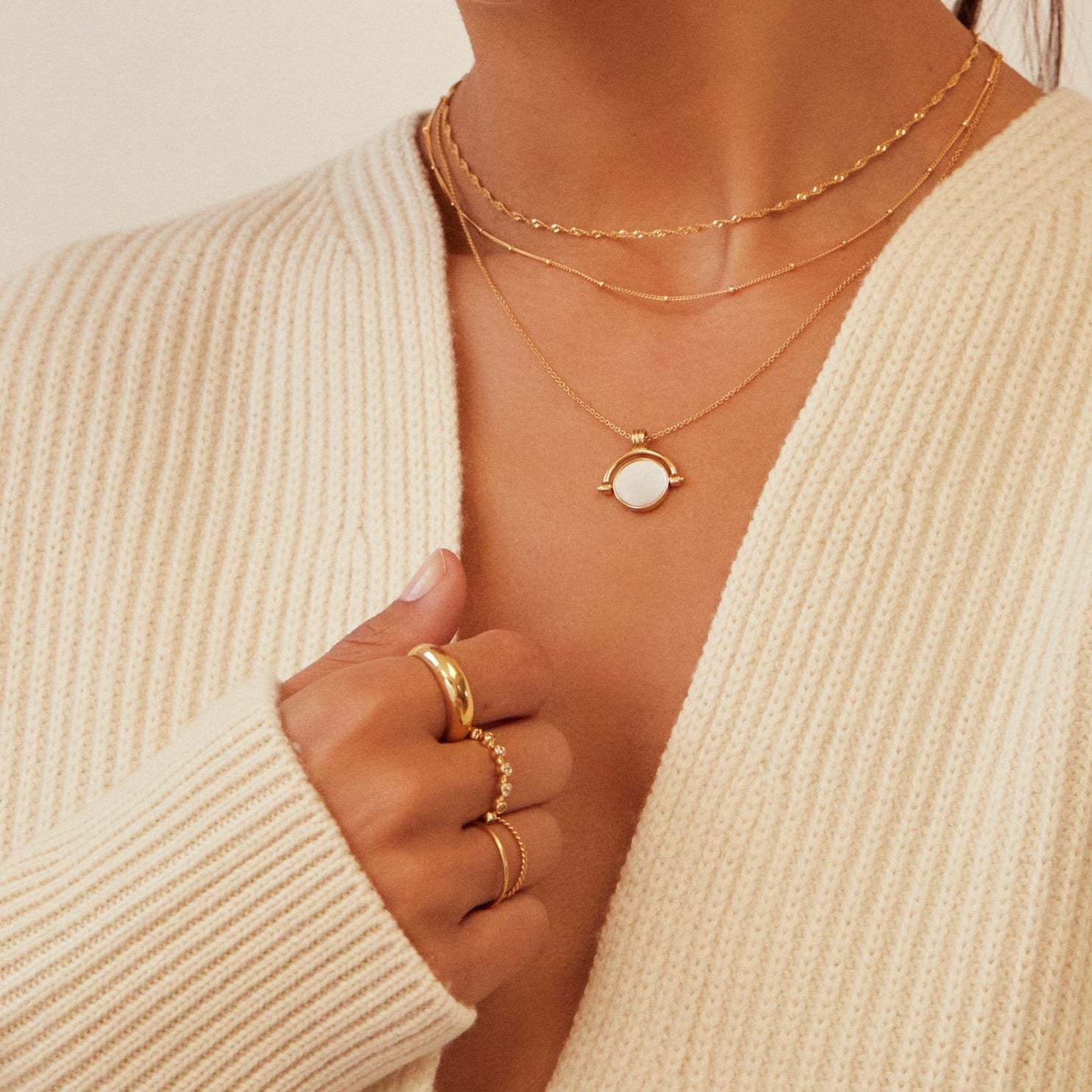 Close up of a woman wearing a gold mother of pearl spinning disc necklace layered with a gold twisted rope necklace, gold plaine dome ring and other gold rings on her fingers and a cream knitted jumper