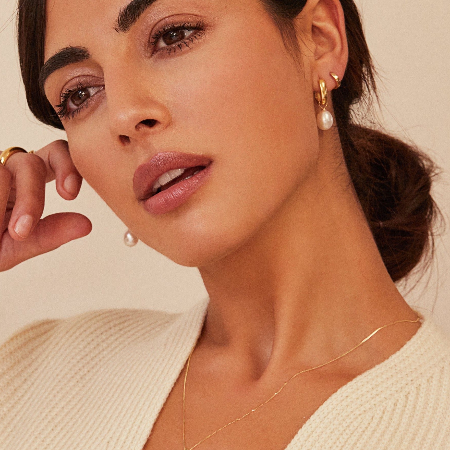 Gold thick squared hoop pearl drop earrings in the ears of a brunette woman wearing a cream knitted jumper with her head tilted 