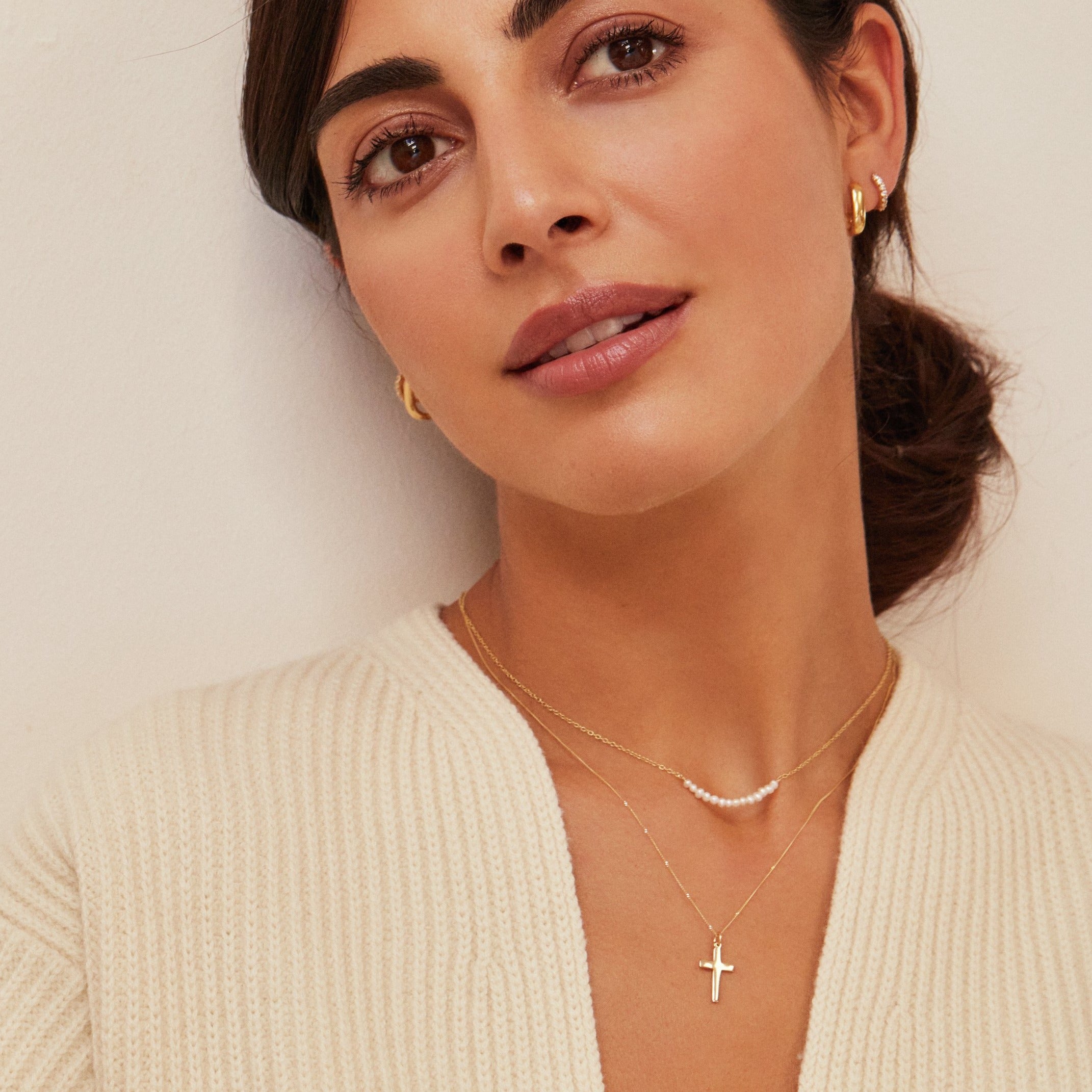 Brunette woman wearing a gold small pearl cluster choker around her neck layered with a gold cross pendant necklace and gold thick squared hoop earrings in her ears