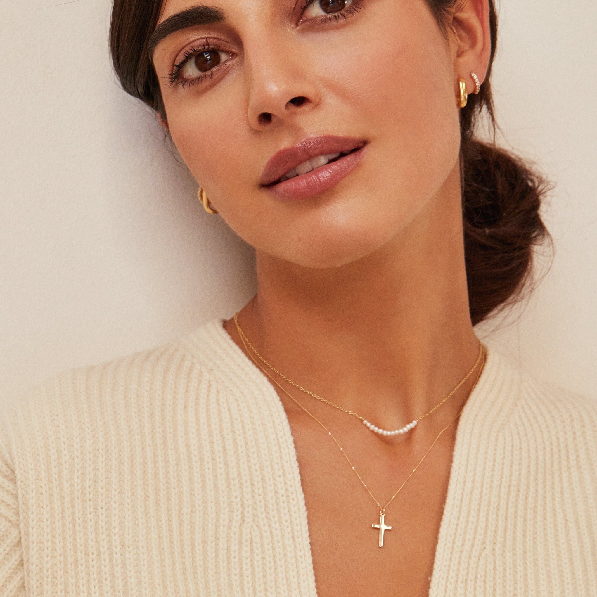A woman wearing a gold small pearl cluster choker layered with a gold cross pendant necklace around her neck and gold thick squared hoop earrings in her ear lobes