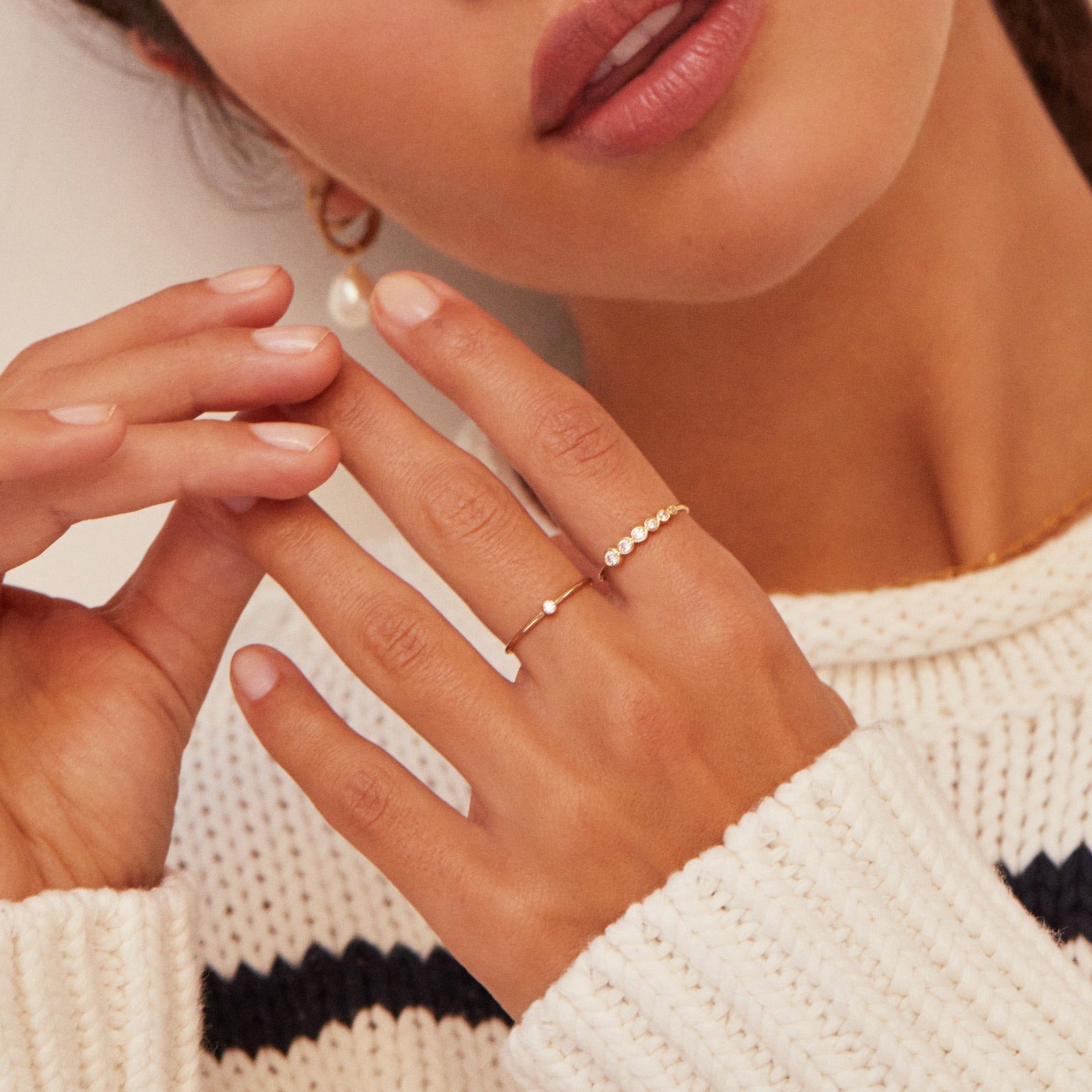 Gold thin diamond style stacking ring on one finger and a gold graduated diamond style stacking ring on another finger of a brunette woman wearing a striped cream and black jumper