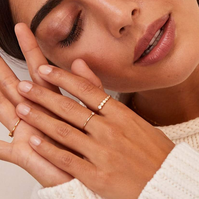 Close up of gold graduated diamond style stacking ring on a finger with a diamond ring on a another finger of a brunette woman with her head to the side wearing a cream jumper