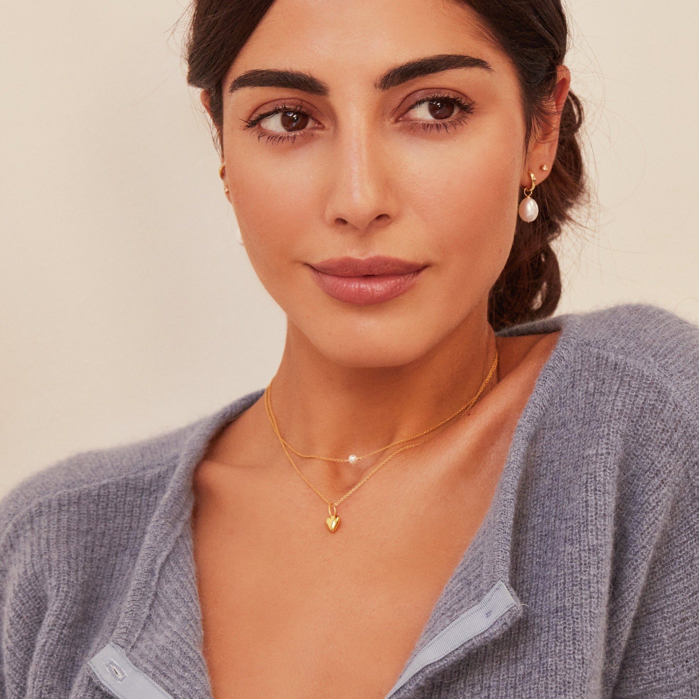 Gold heart pendant necklace around a brunette woman's neck layered with a diamond necklace also wearing a pearl earring and gold small diamond stud in her ear lobe and a grey knitted top