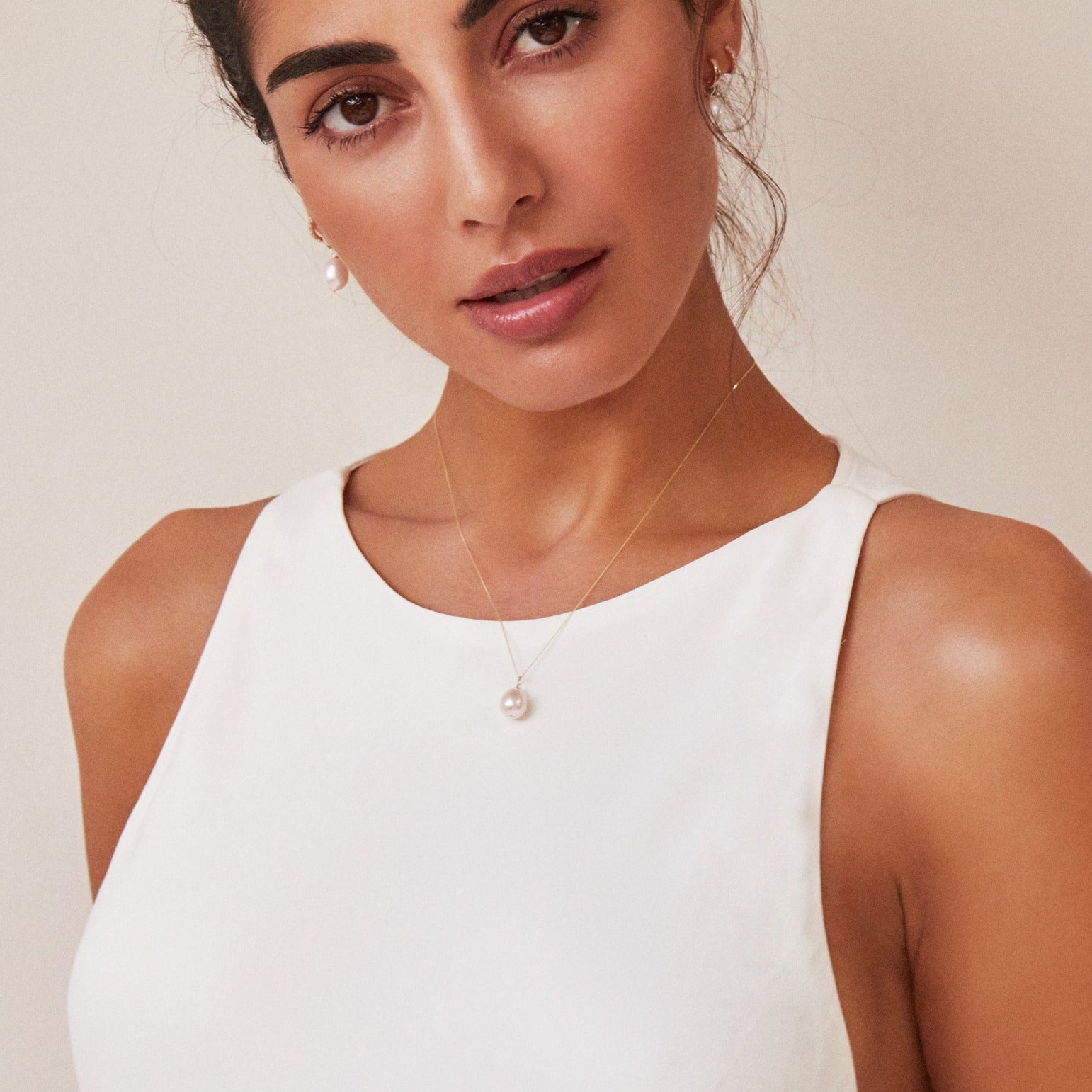 Gold large single pearl necklace around neck with white high neck top on a brunette woman wearing gold plain huggie pearl drop hoop earrings in her ears