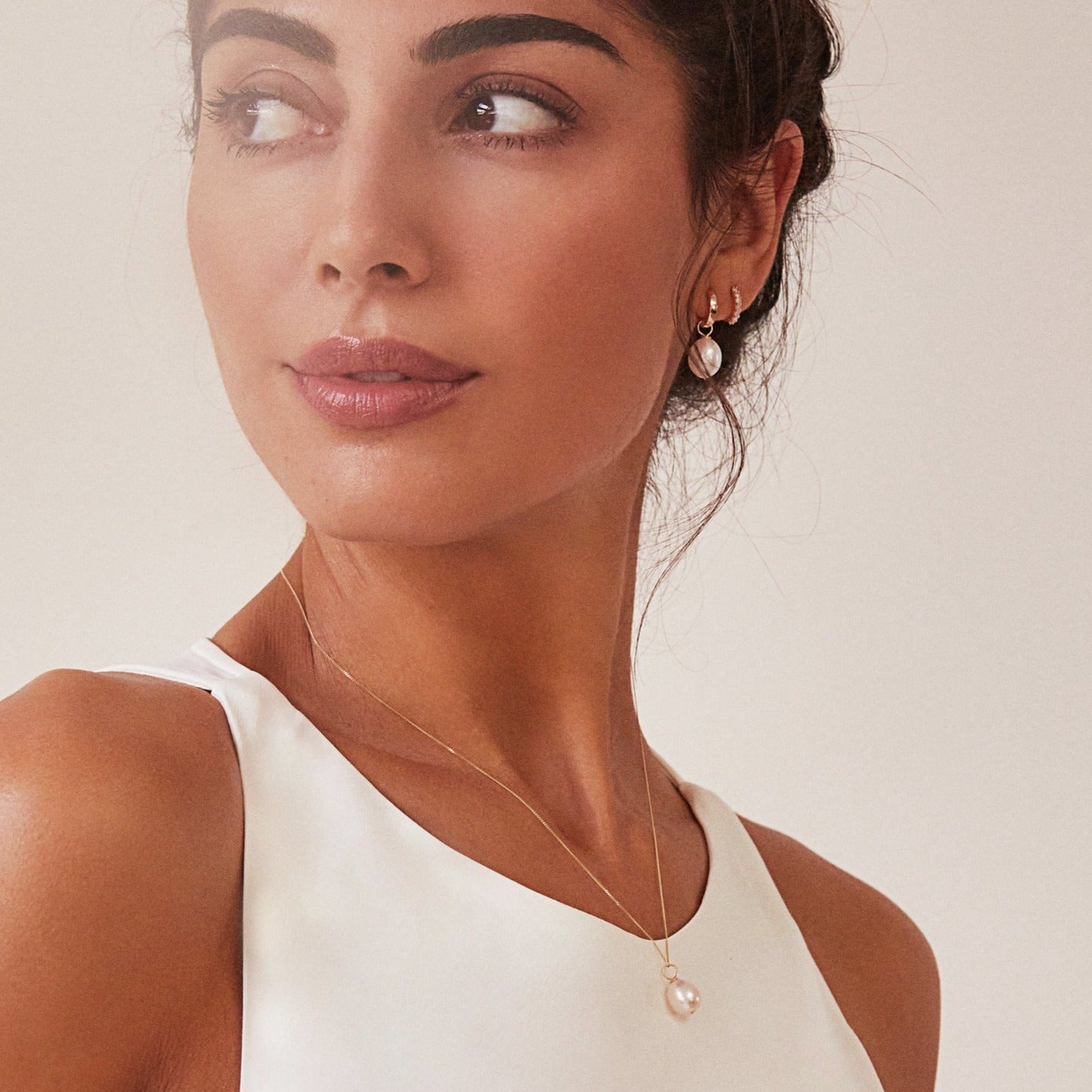 Gold large single pearl necklace around neck with white high neck top on a brunette woman looking to the side wearing a gold plain huggie pearl drop hoop earring in her ear lobe