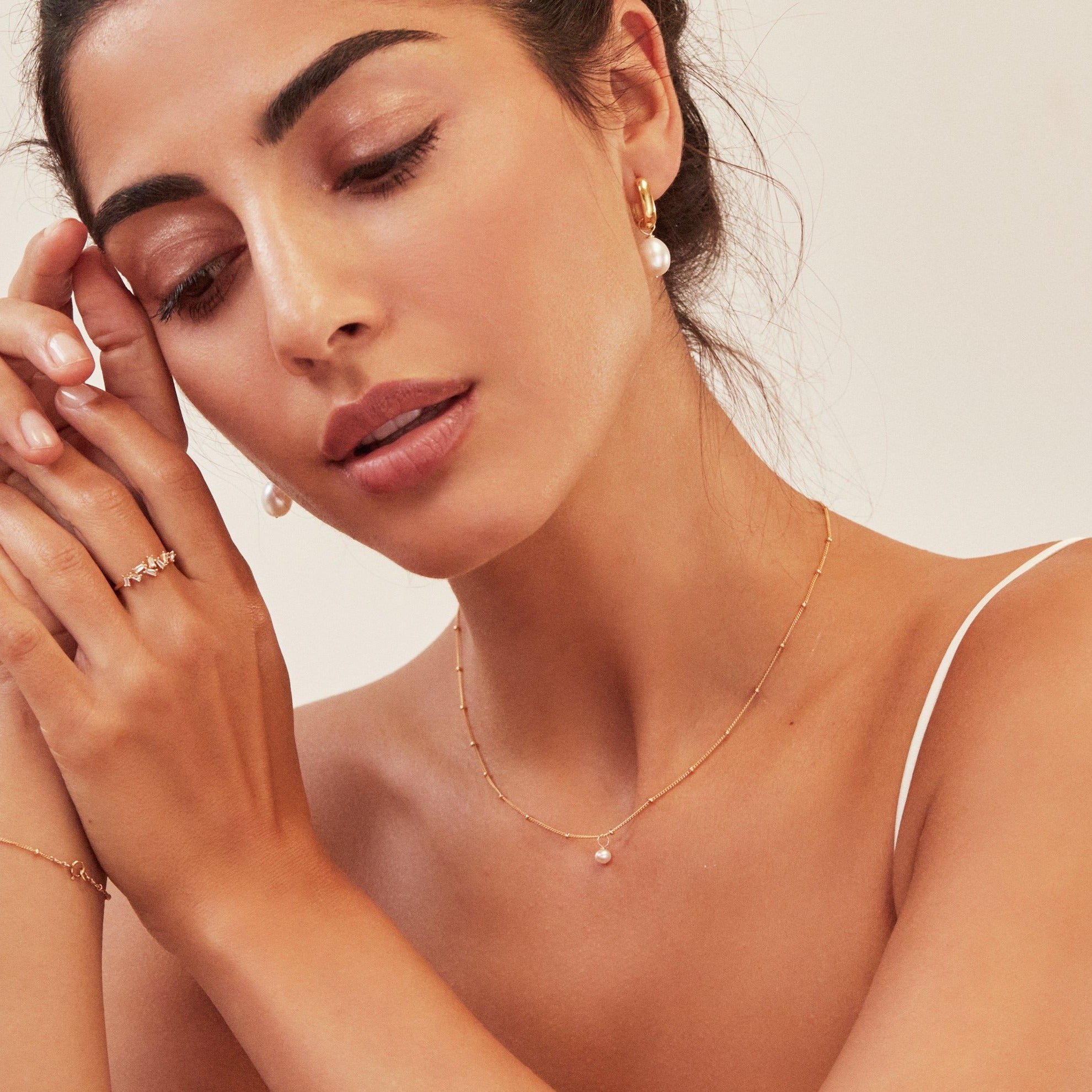 Woman wearing a gold single pearl satellite necklace around her neck, gold thick squared hoop pearl drop earrings in her ears and a gold diamond style baguette ring on her finger clasping her hands together next to her face
