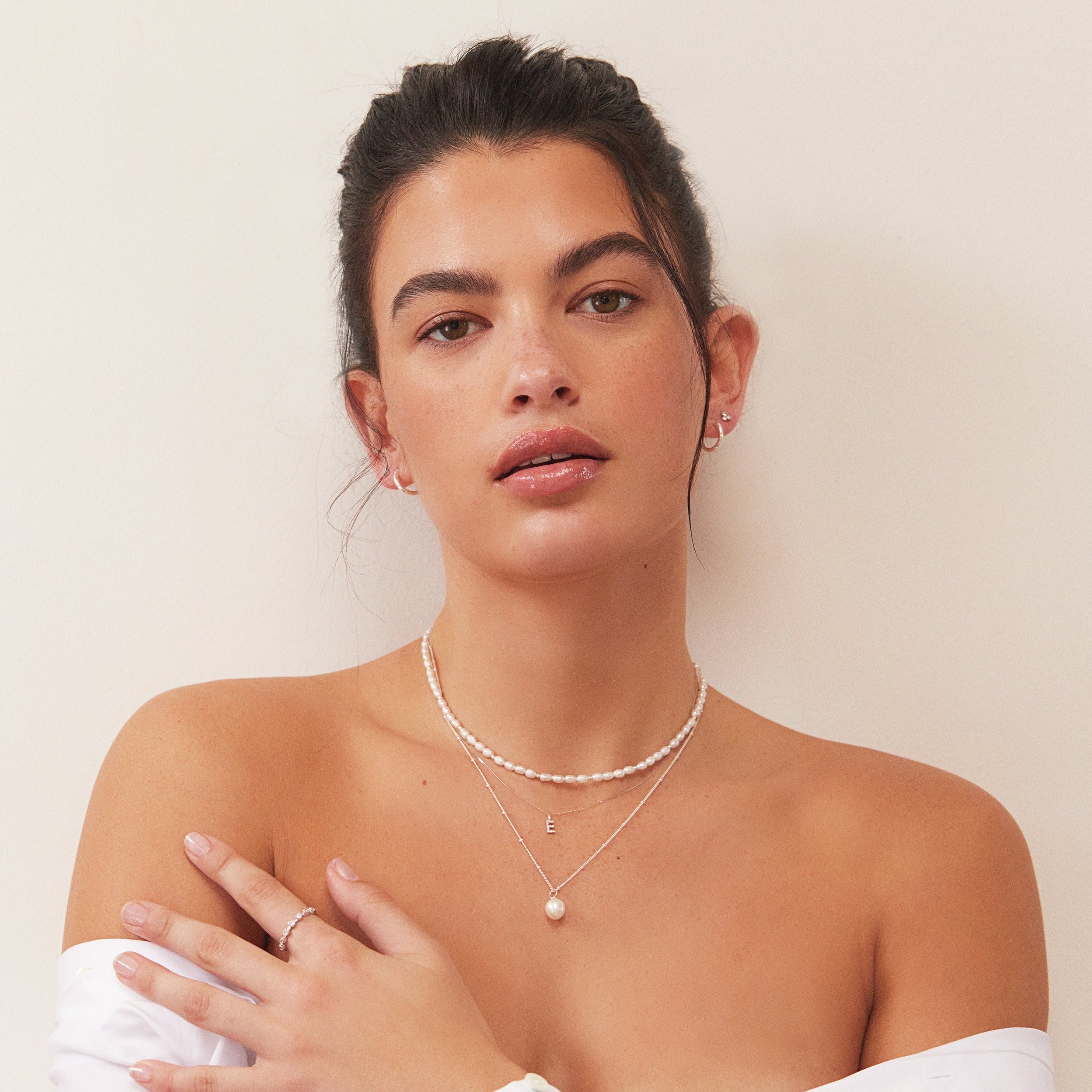 A brunette woman a silver beaded seed pearl choker layered with a silver 'E' letter necklace and a silver satelite single pearl choker around her neck and a off-the-shoulder white shirt