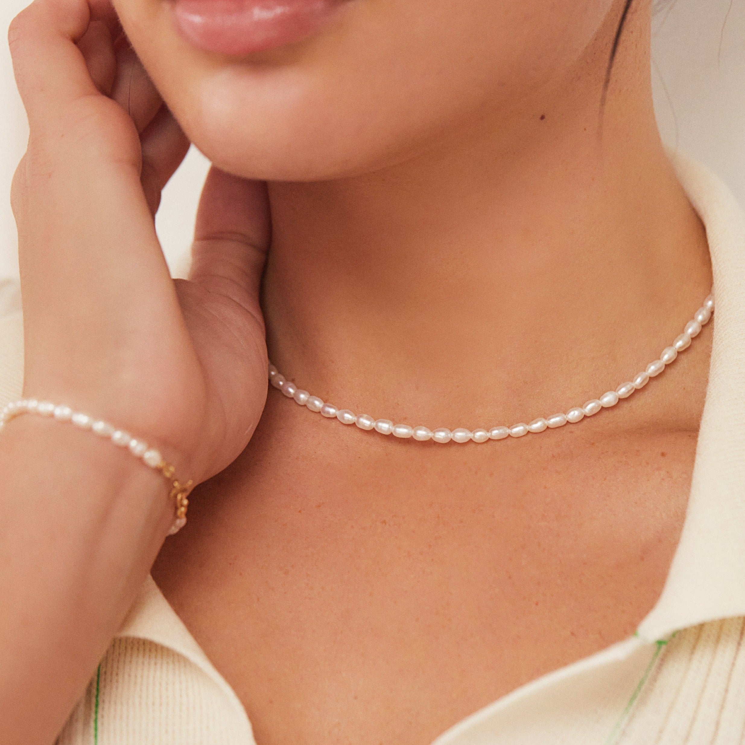 Close up of a woman wearing a gold seed pearl choker around her neck and a gold seed pearl bracelet around her wrist with her hand on her face