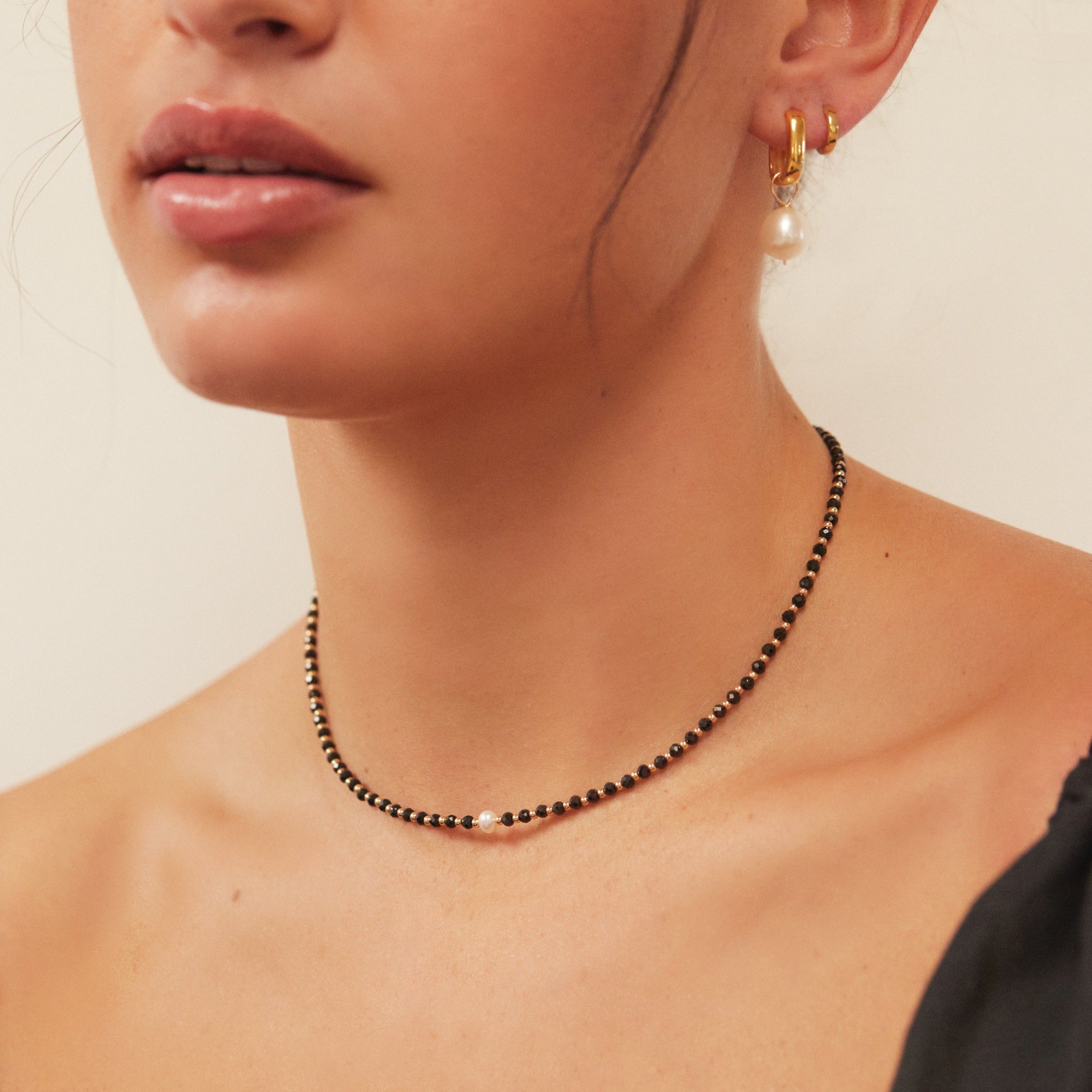 Close up shot of a gold spinel gemstone choker around the neck of a woman wearing a gold thick squared hoop pearl drop earring in her ear