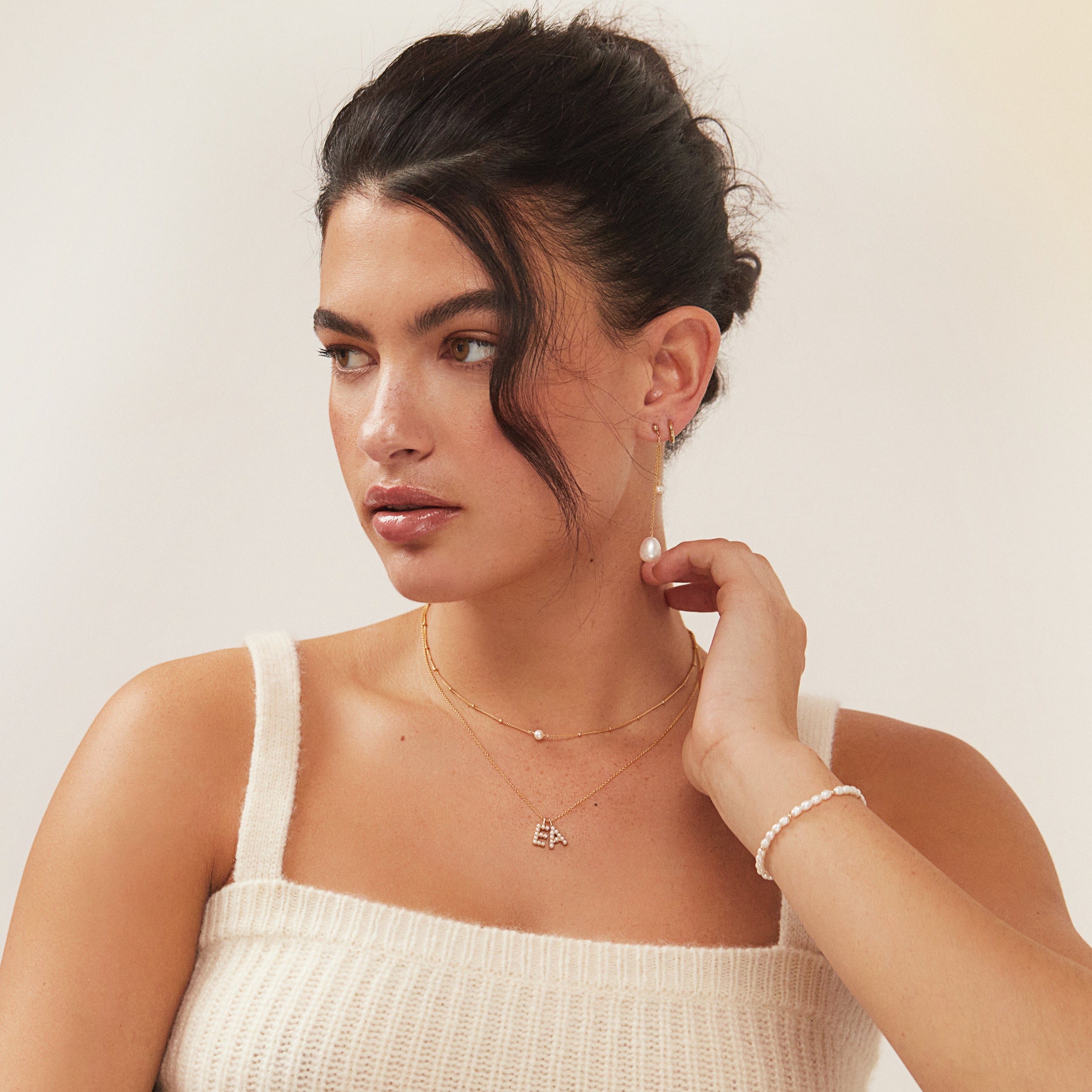 Gold satellite single pearl choker and an 'E' and 'A' inital necklace around a neck of a woman also wearing gold beaded seed pearl bracelet on her wrist and a gold large pearl drop ear threader in her ear lobe