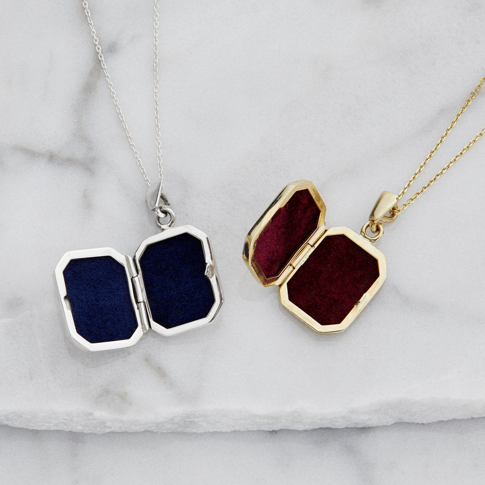 An open silver small square locket necklace with blue velvet lining next to an open gold small square locket necklace with red velvet lining on a marble surface