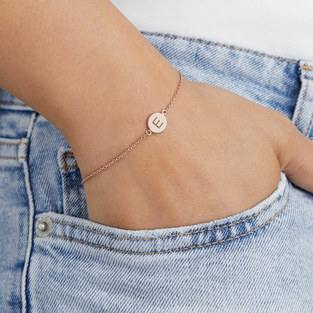 Rose gold personalised disc bracelet with the letter 'E' engraved on a wrist with the hand in blue denim pockets