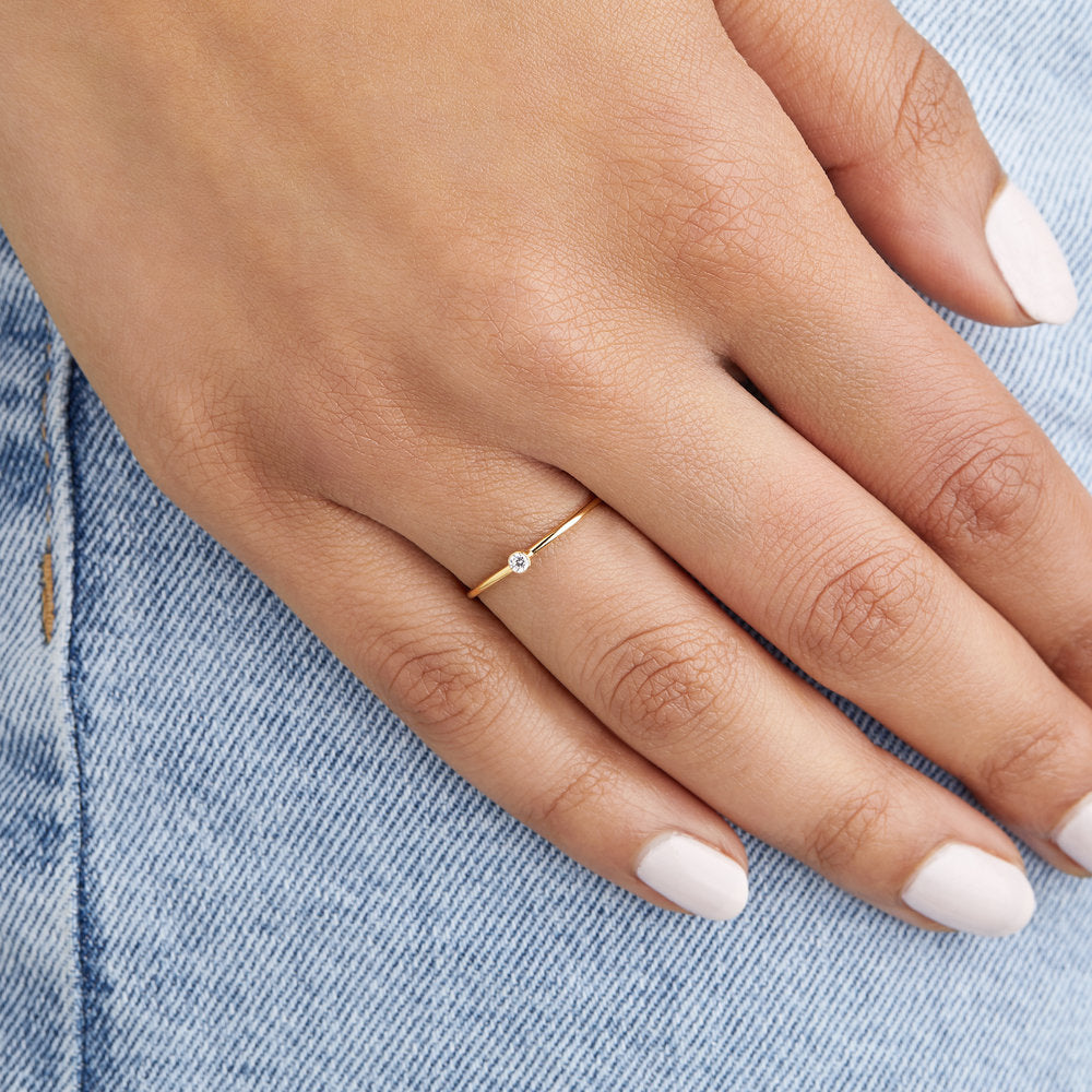 Gold thin diamond style stacking ring on a finger of a hand placed over light blue denim