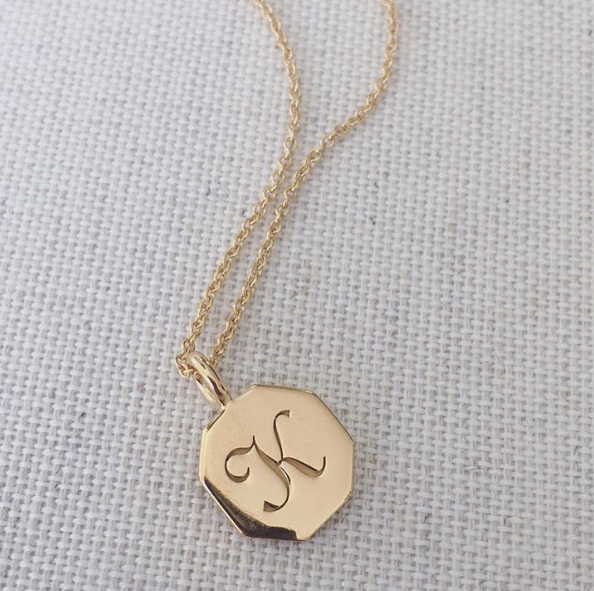 Gold personalised hexagon necklace with the letter 'K' engraved on it on a white woven surface