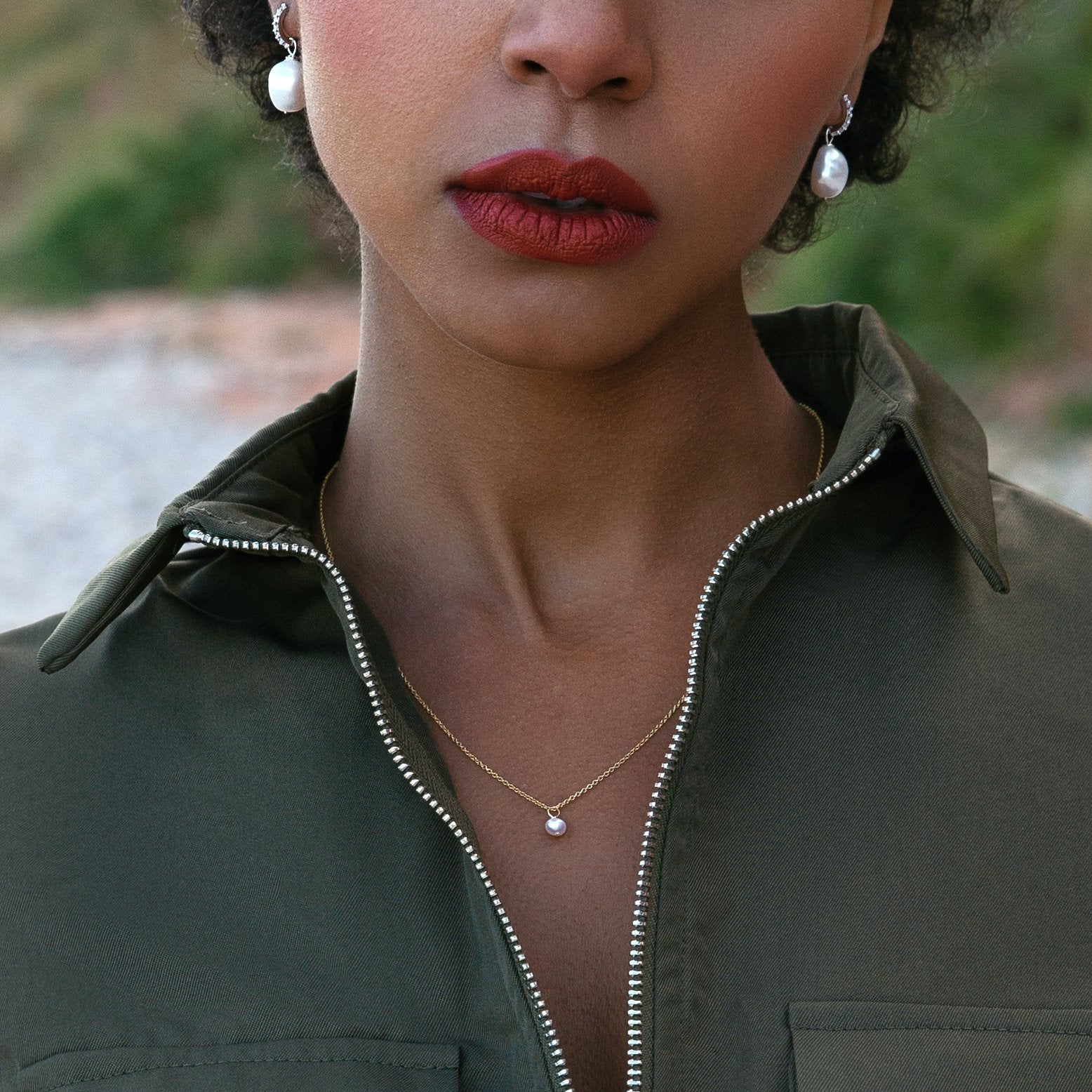 Woman with red lipstick on her lips wearing a gold single pearl necklace around her neck, silver huggie hoop pearl drop earrings in her ears and a army green top with a zip collar