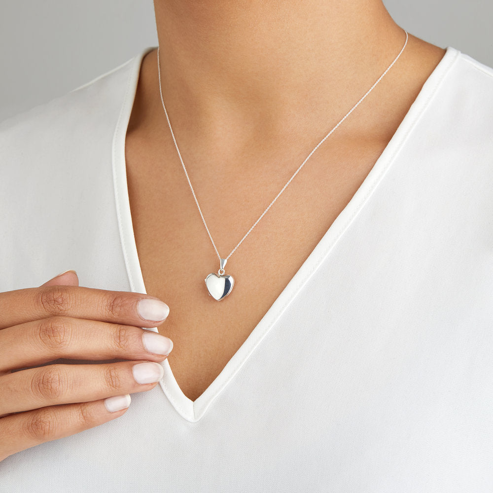 Silver small heart locket necklace around the neck of a woman wearing a white V neck top