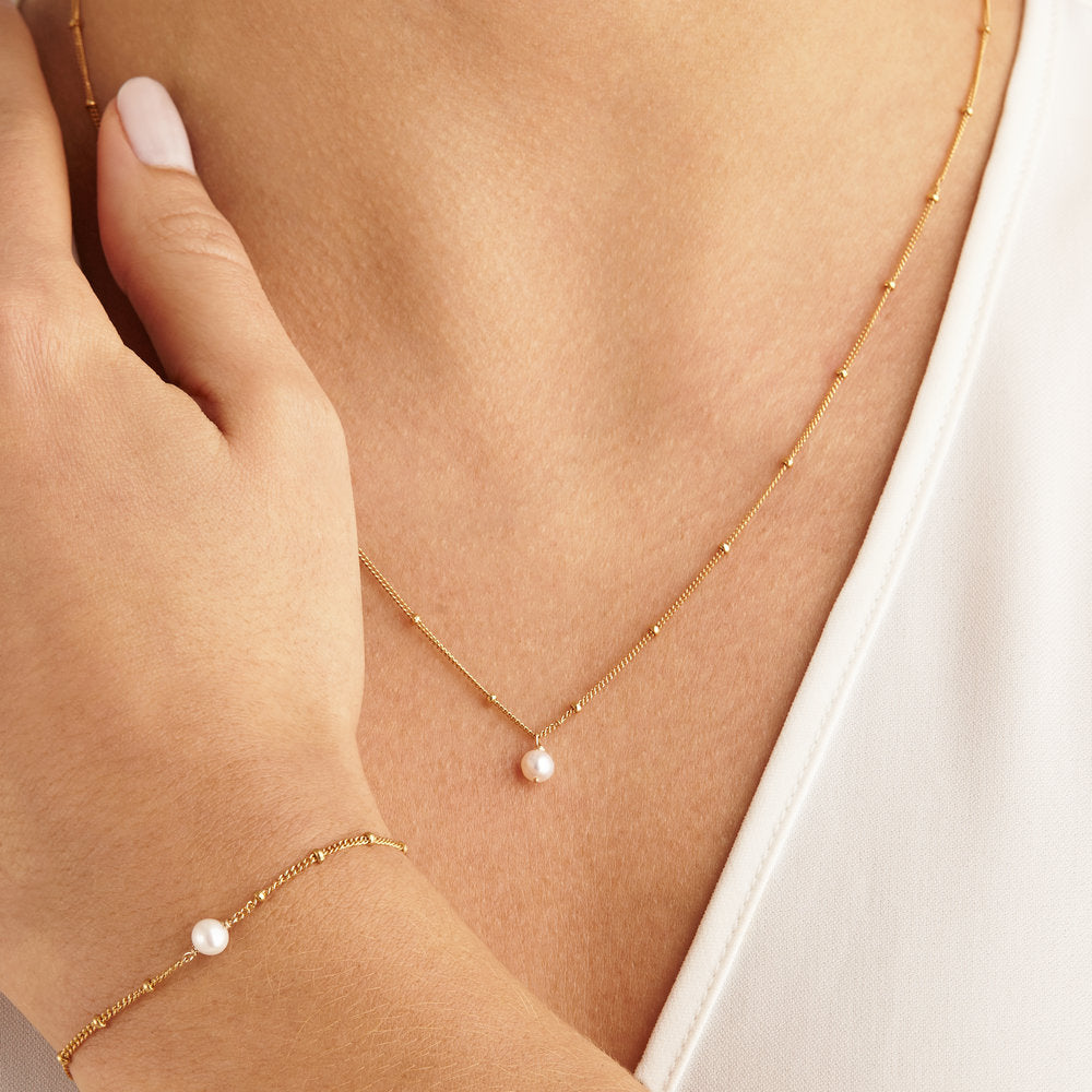 Gold single pearl satellite necklace around a neck of a woman wearing a white V-neck top and a gold single pearl satellite bracelet