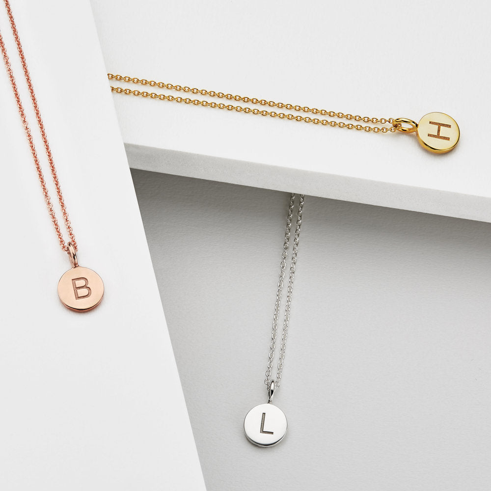 Silver small personalised disc necklace 'L', rose gold small personalised disc necklace 'B' and gold small personalised disc necklace 'H' on white surfaces
