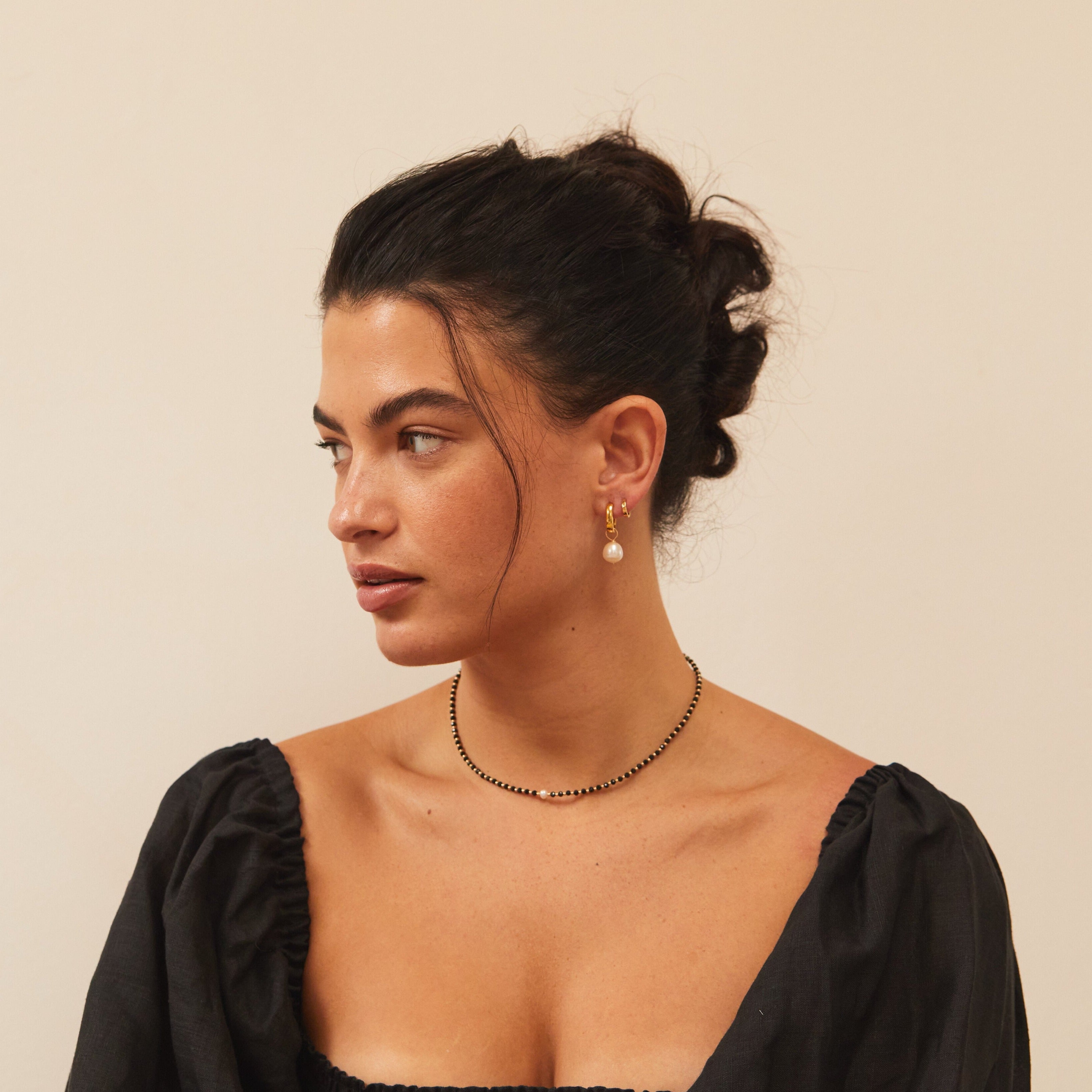 Brunette woman with black sleeves looking to the side wearing a gold spinel gemstone choker around her neck and a gold huggie pearl drop earring in one ear 